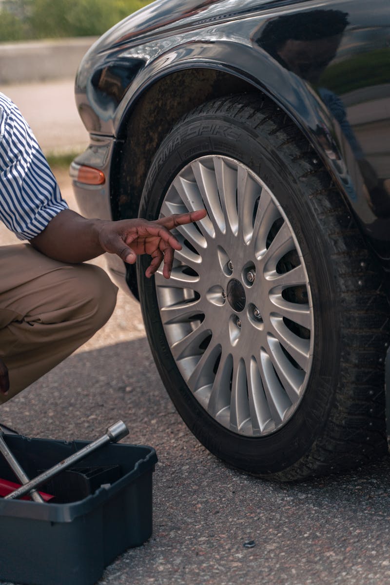 An adult male pointing at a car tire, suggesting a focus on vehicle maintenance or inspection outdoors.